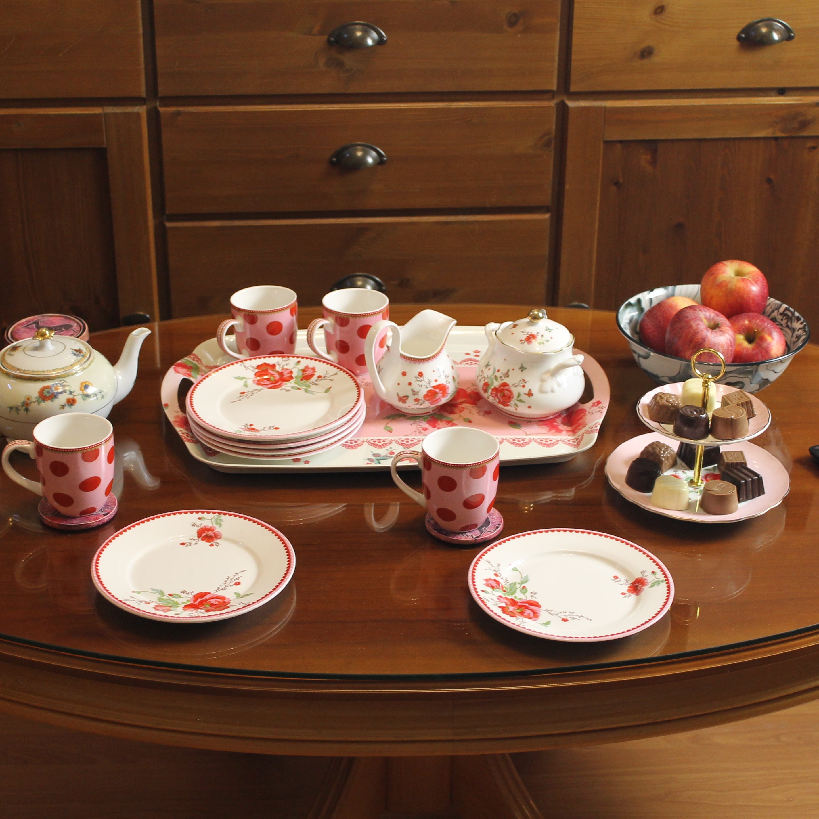Tea set with plates, cups, and saucers on a glass-top table with wooden cabinets in the background.